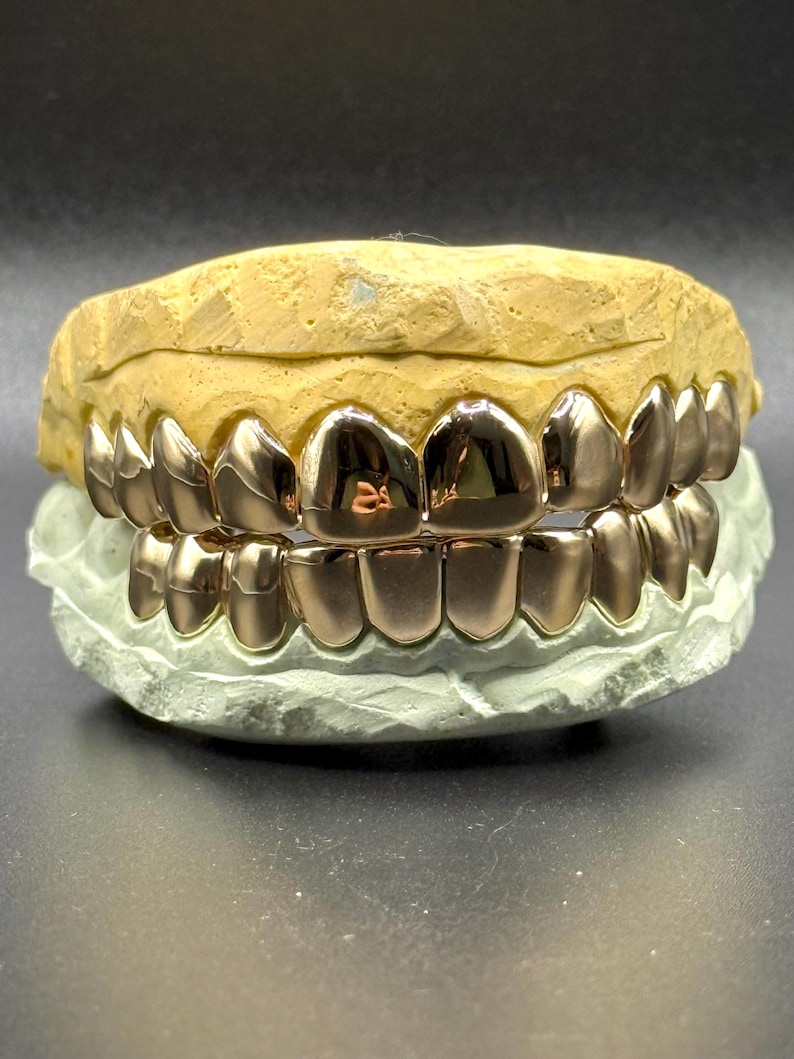 Puede incluir: Un juego de parrillas dentales de color oro rosa pulido ajustadas sobre un molde dental beige y verde claro. Las parrillas met&aacute;licas cubren los dientes superiores e inferiores, con una superficie lisa y reflectante que capta la luz en cada pieza.