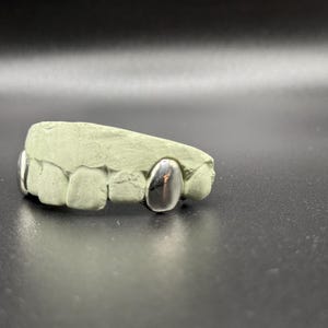 Puede incluir: Un molde dental con tres fundas de dientes plateadas. El molde es de color verde claro y est&aacute; sobre una superficie oscura. Las fundas son lisas y reflectantes, contrastando con la textura mate del molde.