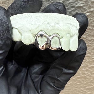 May include: Silver-colored dental grill with a heart-shaped design on one tooth and a smooth surface on the other. The grill is set on a light green dental mold, held by a person wearing black gloves. The background is a neutral color.
