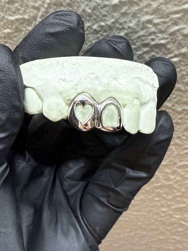 May include: Silver-colored dental grill with a heart-shaped design on one tooth and a smooth surface on the other. The grill is set on a light green dental mold, held by a person wearing black gloves. The background is a neutral color.