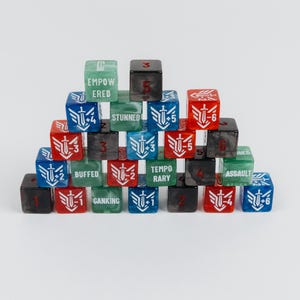 Puede incluir: Una pirámide de dados de juego de seis caras multicolores en rojo, azul, verde y negro. Cada dado presenta texto impreso en blanco que incluye palabras como EMPOWERED, STUNNED, BUFFED, TEMPORARY, GANKING y ASSAULT, junto a iconos de espada y números.