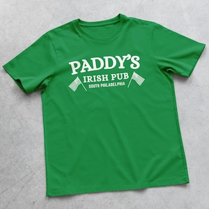 Puede incluir: Camiseta verde con texto blanco que dice "Paddy's Irish Pub South Philadelphia" con dos banderas americanas blancas a cada lado del texto.