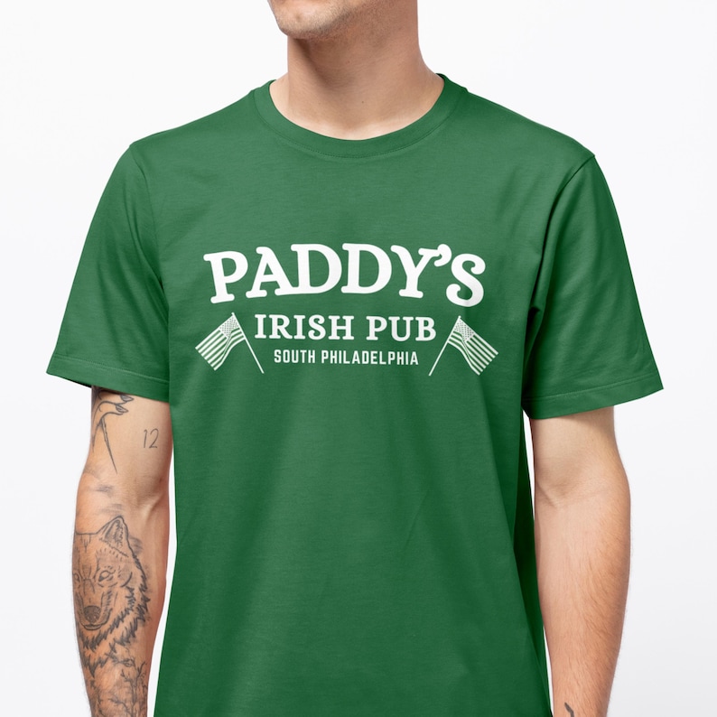 Puede incluir: Una camiseta verde con texto blanco que dice "Paddy's Irish Pub South Philadelphia" con dos banderas americanas blancas a cada lado del texto.