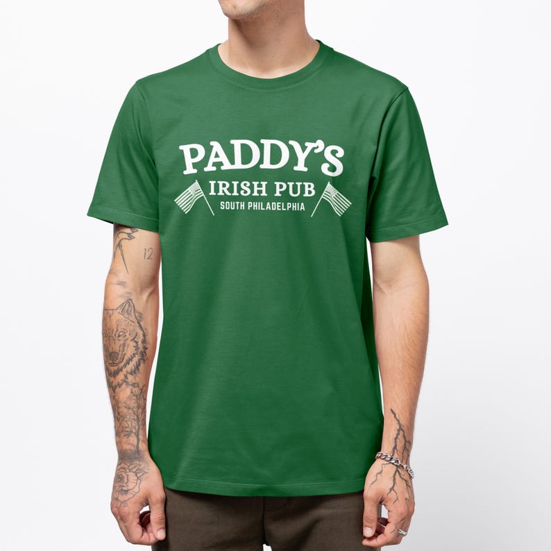 Puede incluir: Camiseta verde con texto blanco que dice "Paddy's Irish Pub South Philadelphia" con dos banderas americanas blancas a cada lado del texto.