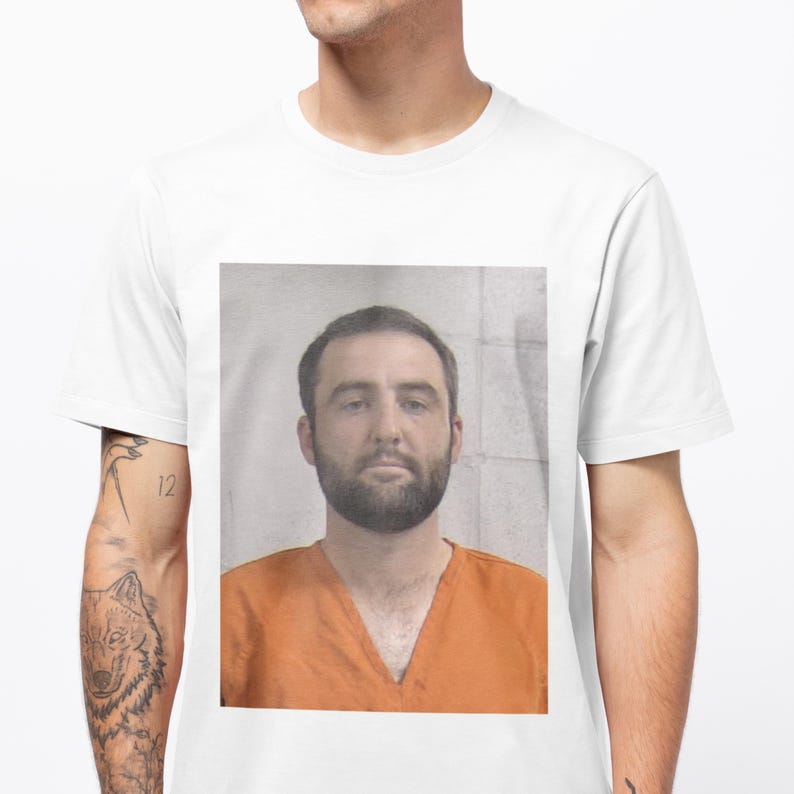 May include: A white t-shirt with a printed image of a man in an orange jumpsuit. The man has short brown hair, a beard, and a serious expression.