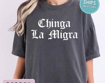 May include: A dark gray t-shirt with the text "Chinga La Migra" in white gothic font. The shirt has a crew neck and short sleeves. The word "Pepper" is in a pink rectangle at the bottom of the shirt.