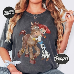 May include: Dark gray Comfort Colors t-shirt featuring a cartoon illustration of a horse and a cowgirl hugging. The cowgirl has red hair and is wearing a white shirt, blue jeans, and cow-print chaps. The horse is brown. The text "Pepper" is on the bottom right.
