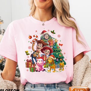 A woman wearing a pink t-shirt with a Christmas-themed graphic featuring characters from the Toy Story movie, including Woody, Buzz Lightyear, and other beloved characters.