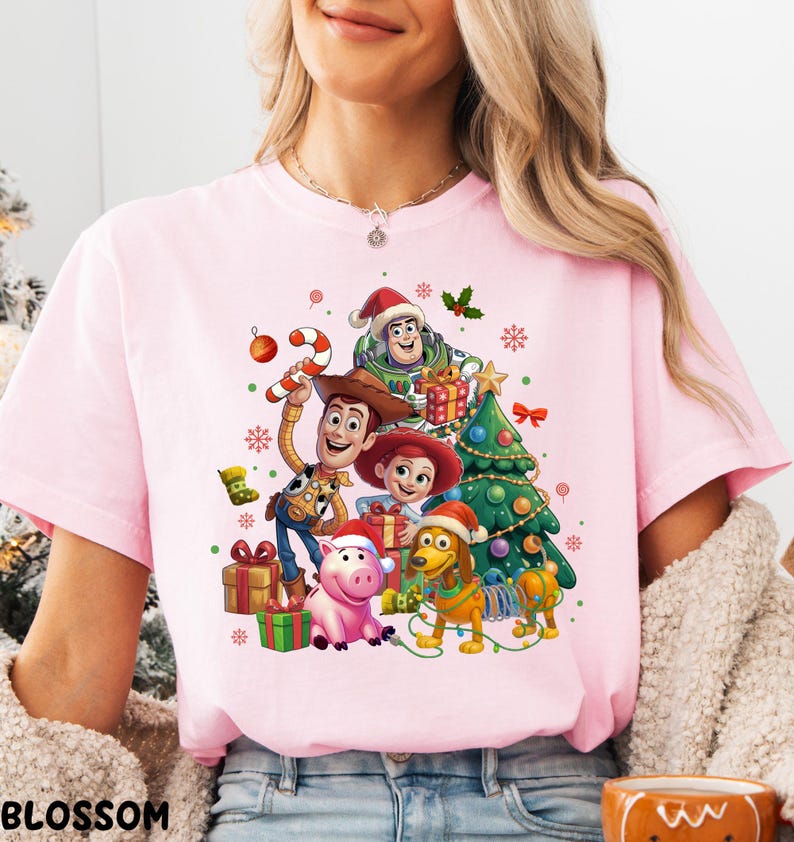 A woman wearing a pink t-shirt with a Christmas-themed graphic featuring characters from the Toy Story movie, including Woody, Buzz Lightyear, and other beloved characters.