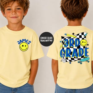 May include: A light yellow t-shirt featuring a small blue smiley face graphic with the name James on the front. The back displays a large black and white checkered flag graphic with the text 3rd Grade, a pencil, a skateboard, and a skeleton hand.