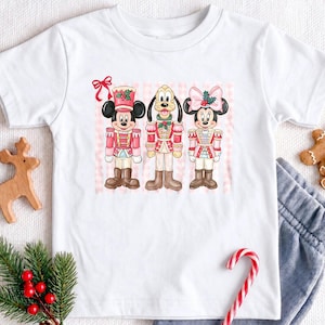 May include: A white short-sleeve t-shirt featuring a graphic of Mickey Mouse, Pluto, and Minnie Mouse dressed as nutcrackers. The characters are wearing red and pink uniforms with brown boots, set against a light pink striped background.
