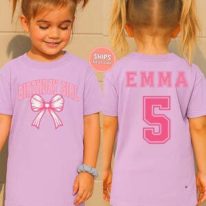A young girl wearing a purple t-shirt with the text "BIRTHDAY GIRL" and a pink bow on the front.