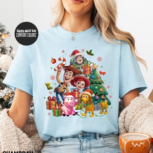 A woman wearing a light blue t-shirt with a colorful graphic of characters from the Toy Story movie, including Woody, Buzz Lightyear, and a pig, surrounded by Christmas decorations.