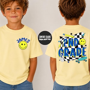 May include: A pale yellow t-shirt featuring a front graphic with a blue smiley face and the name James. The back graphic displays a black and white checkered flag, a yellow pencil, a skateboard, and the blue text 2nd Grade with a skeleton hand.