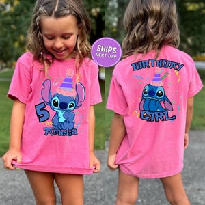May include: A pink t-shirt featuring a blue cartoon character wearing a party hat, with the number 5 and the name "Amelia" printed on the front. The back of the shirt has the words "BIRTHDAY GIRL" and the same cartoon character.