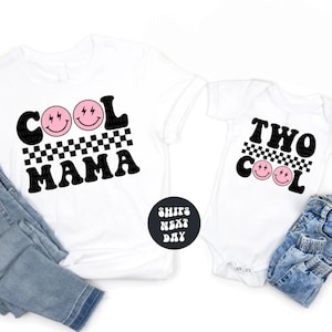 May include: White t-shirt and baby onesie with matching "Cool Mama" and "Two Cool" designs. Each features a black and white checkered pattern, pink smiley faces, and lightning bolt accents. Denim jeans are visible.