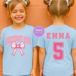 A young girl wearing a light blue t-shirt with a pink bow and the text "BIRTHDAY GIRL" printed on it, smiling at the camera.