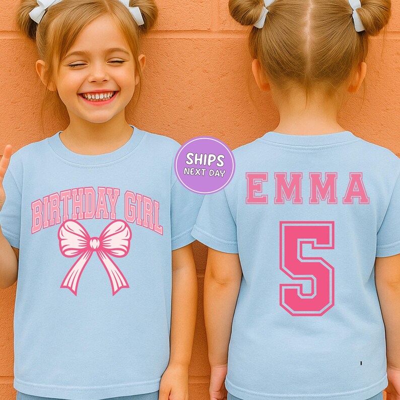 A young girl wearing a light blue t-shirt with a pink bow and the text "BIRTHDAY GIRL" printed on it, smiling at the camera.