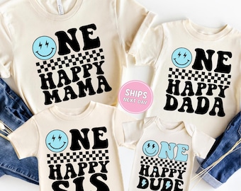 May include: Four cream-colored t-shirts and a onesie with matching designs. Each shirt features the text "ONE HAPPY" above a checkered pattern, followed by "MAMA", "DADA", "SIS", and "DUDE". A light blue smiley face and lightning bolts complete the design.