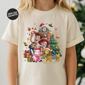 A t-shirt featuring a colorful illustration of characters from the Toy Story movie, including Woody, Buzz Lightyear, and other beloved characters, surrounded by Christmas decorations.