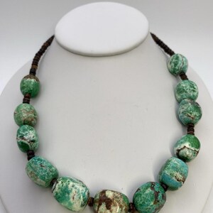 May include: A green and brown stone bead necklace with a brown cord. The beads are oval shaped and have a mottled pattern.