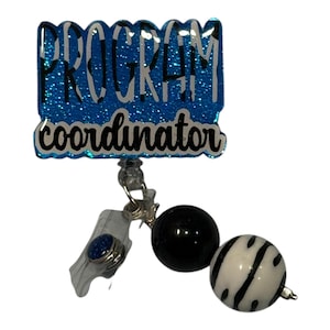May include: A blue glitter badge reel with the words "PROGRAM coordinator" in white lettering. The reel has a clear plastic clip, a black bead, and a black and white striped bead.