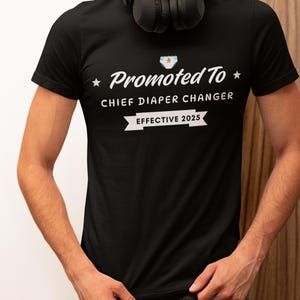 Puede incluir: Camiseta negra con texto blanco que dice "Promocionado a Jefe de Cambio de Pañales Efectivo 2025". Un pequeño pañal blanco con un diseño azul y blanco está por encima del texto.