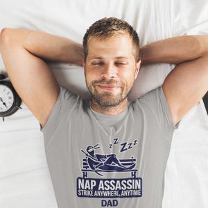 May include: A gray t-shirt with a blue graphic of a sleeping ninja on a bench. The text on the shirt reads "Nap Assassin Strike Anywhere, Anytime Dad".