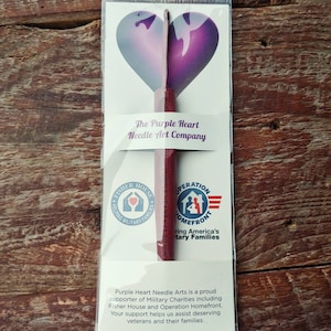 May include: A purple heart-shaped crochet hook with a wooden handle. The packaging includes the text "The Purple Heart Needle Art Company" and logos for Fisher House and Operation Homefront. The text reads "Purple Heart Needle Arts is a proud supporter of Military Charities including Fisher House and Operation Homefront. Your support helps us assist deserving veterans and their families."