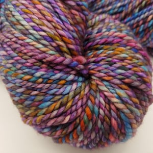 May include: Close-up of a skein of colorful yarn. The yarn is a blend of purple, blue, orange, and green hues, creating a vibrant and textured appearance. The thick, twisted strands suggest a chunky knit or crochet project. Ideal for fiber art.