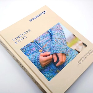 May include: A book titled "Timeless Knits" by Malabrigo. The cover features a close-up of a blue, purple, and pink knitted cardigan. The book's cover is a light beige color with blue text. The book is about classic knitting patterns from Uruguay.