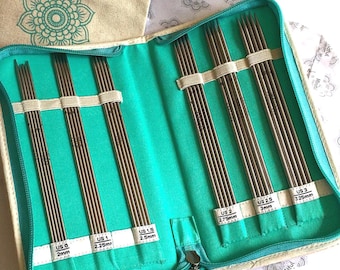 Op de afbeelding: Een set dubbelpuntige breinaalden van roestvrij staal in een turquoise stoffen etui. De naalden zijn gelabeld met de maten US 0 (2mm), US 1 (2,25mm), US 1,5 (2,5mm), US 2 (2,75mm), US 2,5 (3mm) en US 3 (3,25mm) op witte stoffen lipjes.