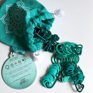May include: A teal canvas bag with a white mandala design contains a set of teal and green stitch markers. The bag is labeled "Knitters Pride" and includes a description of the product. The stitch markers are in various shades of teal and green.