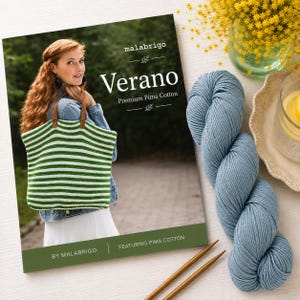 Puede incluir: Un libro de patrones Malabrigo Verano Premium Pima Cotton que muestra un bolso de ganchillo a rayas verdes y blancas con asas de cuero marr&oacute;n, acompa&ntilde;ado de una madeja de hilo azul y dos agujas de tejer de madera sobre una superficie blanca.