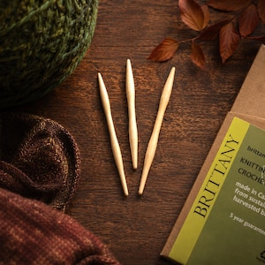 May include: Three light-colored wooden knitting needles are arranged on a dark wooden surface. A ball of green yarn and a brown and burgundy knitted fabric are in the background. A cardboard box with the word "BRITTANY" and text about knitting needles and crochet hooks is also visible.