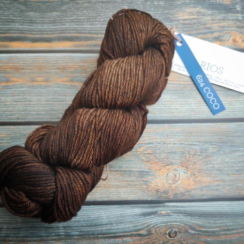 Malabrigo Rios Coco Tonal Espresso Superwash Merino Yarn skein in rich brown color with tag on rustic wooden background, showcasing the yarn's texture and earthy tones, from the Malabrigo brand.