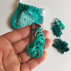 May include: A set of teal colored plastic stitch markers, organized on a metal ring. The markers are arranged in a graduated color scheme, with the darkest teal at the top and the lightest teal at the bottom.