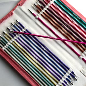 May include: A set of colorful knitting needles in a pink case. The needles are various colors, including purple, green, gold, and pink. Each needle has a silver tip and is labeled with its size in US measurements.