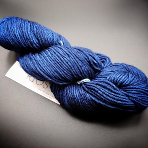 May include: A skein of dark blue yarn with a white label. The yarn is tightly wound and has a textured appearance. The label reads "KIOS". The yarn is likely used for knitting or crocheting projects.