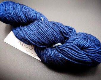 May include: A skein of dark blue yarn with a white label. The yarn is tightly wound and has a textured appearance. The label reads "KIOS". The yarn is likely used for knitting or crocheting projects.