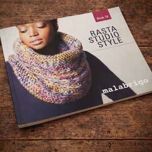 May include: A book titled "Rasta Studio Style" with the word "malabrigo" on the cover. The cover features a woman wearing a colorful knitted cowl. The cowl is a mix of pink, yellow, and purple hues. The book is on a wooden surface.