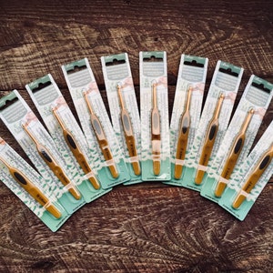 May include: A set of ten gold crochet hooks, packaged individually. Each hook is labeled with its size in millimeters and inches. The hooks are arranged in a fan shape on a wooden surface.