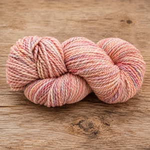 May include: A skein of hand-dyed yarn featuring a variegated color palette of soft peach, coral, and pale pink tones with subtle flecks of lavender and cream throughout the twisted, multi-ply fiber strands of the thick, soft textile material.