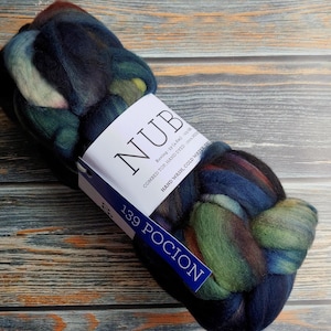 May include: A skein of hand-dyed roving yarn in a blend of colors including blue, green, and brown. The yarn is labeled "NUB" and "139 POCION". The yarn is on a wooden surface.