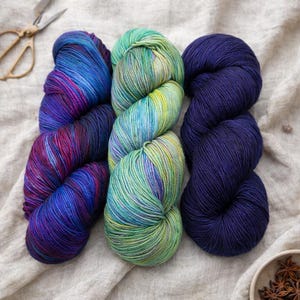 May include: Three skeins of colorful yarn in shades of blue, purple, and green. The yarn is arranged on a neutral-colored fabric, with a pair of scissors in the upper left corner. The yarn appears soft and suitable for knitting or crocheting projects.