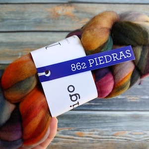 May include: A skein of colorful yarn with a mix of orange, purple, green, and yellow hues. The yarn is labeled with a blue tag that reads "862 PIEDRAS". The yarn is held in a hand.