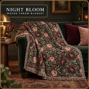 May include: A woven throw blanket featuring a dark background with a dense floral pattern of pink and deep red roses, green leaves, and small berries. The blanket has a fringed edge and the text NIGHT BLOOM WOVEN THROW BLANKET is visible.