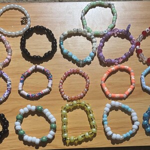 May include: A collection of 16 colourful beaded bracelets, featuring various patterns and colours, including pink, blue, green, white, black, and red.