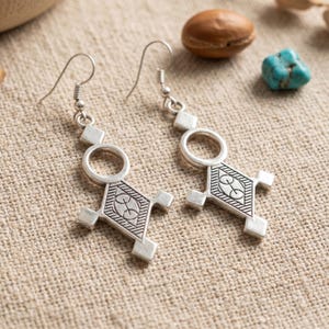 Peut inclure: Une paire de boucles d'oreilles pendantes argentées au design géométrique, composées d'un anneau circulaire relié à une base en forme de losange. La section en losange présente des motifs de lignes gravées et de petites saillies carrées.