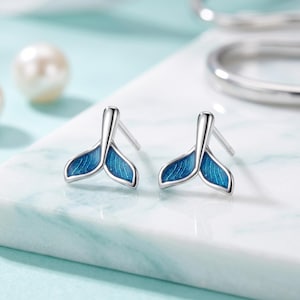 Peut inclure: Une paire de boucles d'oreilles à tige en forme de queue de baleine. Chaque boucle présente un cadre en argent poli entourant un émail bleu vif avec des détails texturés fins qui imitent l'apparence des motifs naturels des nageoires.